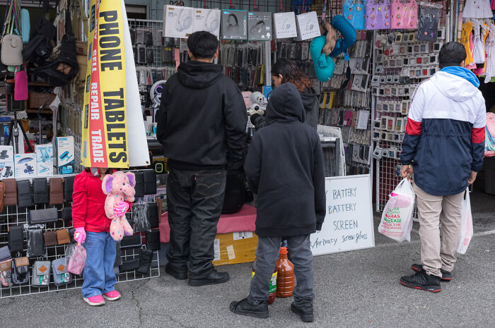 Street photographer captures humor and chaos in everyday life with candid shots of people at a busy market stall.