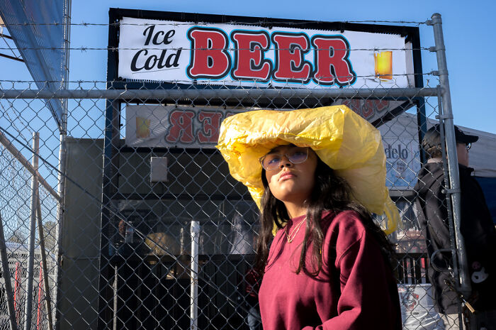Street photographer captures humor and chaos of everyday life with a woman wearing a bright yellow hat near a beer sign.