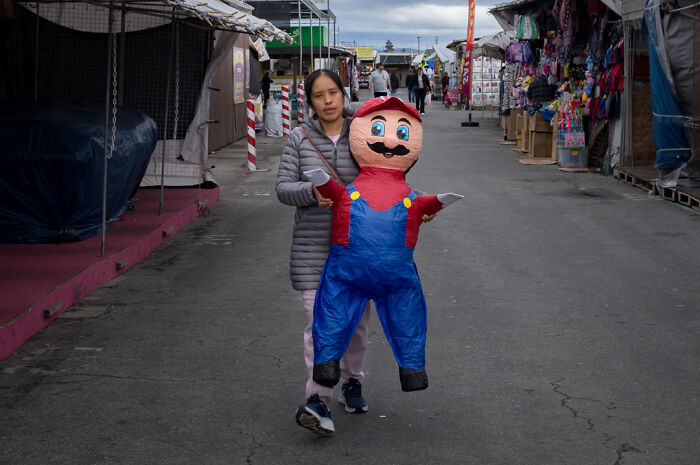 Woman carrying a colorful piñata in a street market, capturing humor and chaos in everyday life by street photographer.