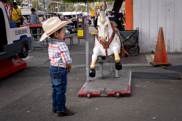Young boy in a cowboy hat observes a mechanical horse at a street scene, capturing humor and chaos in everyday life.