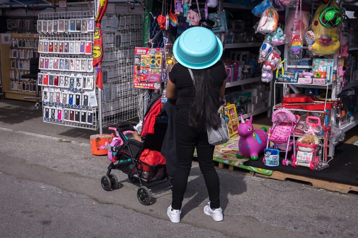 Street photographer captures humor and chaos in everyday life with a woman wearing a basin on her head at a busy outdoor market.