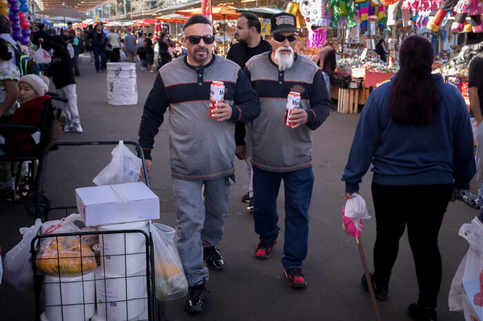 Two men walking with drinks through a busy market, capturing humor and chaos in everyday street life.