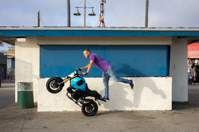 Street photographer captures humor and chaos as a man performs a motorcycle stunt in an urban everyday setting.