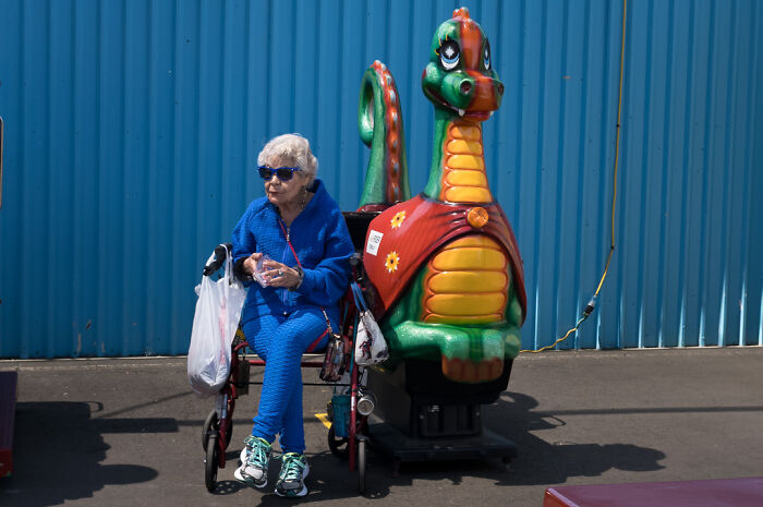 Elderly woman in blue sitting on a walker next to a colorful dragon ride, capturing humor and chaos in everyday street life.