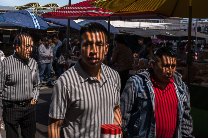 Three men walking in a crowded street market with striped shadows on their faces captured by a street photographer.