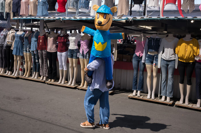 Street photographer captures humor and chaos in everyday life with a person carrying a large bear figure at a market.