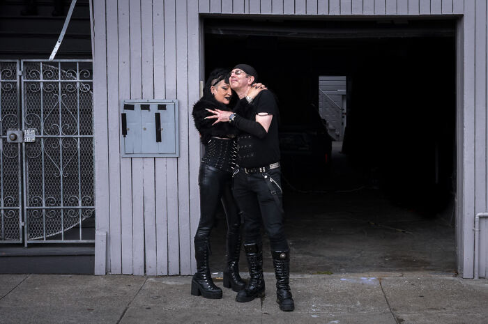 Couple dressed in black leather posing outside a garage, captured by a street photographer spotting humor and chaos in life.