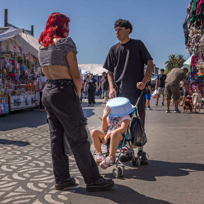 Street photographer captures humor and chaos as a child hides under a basin in a stroller at a busy outdoor market.