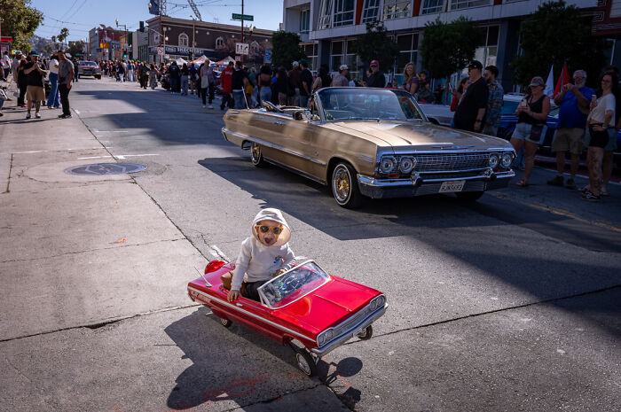 Toddler in a red toy car wearing sunglasses, with a classic convertible and crowd, capturing street photographer humor and chaos.