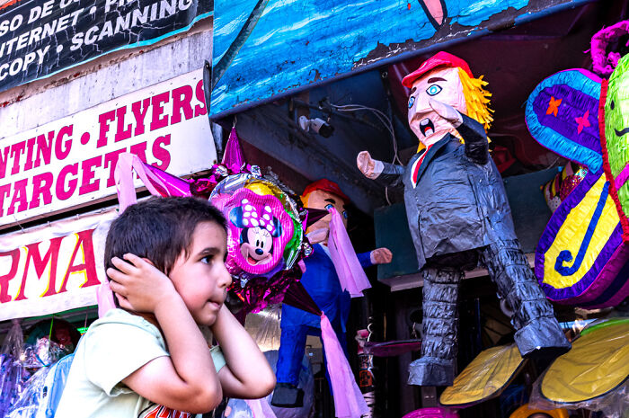 Young boy covering ears near colorful piñatas in busy street scene, capturing humor and chaos in everyday life by street photographer.