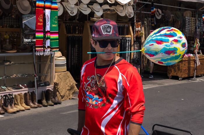 Street photographer captures humor and chaos in everyday life with a man balancing a colorful balloon on a stick in a market setting.