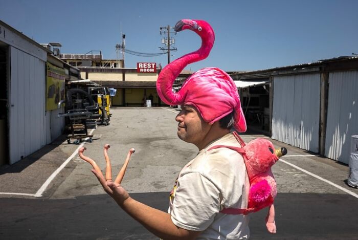 Man wearing a flamingo hat and backpack, holding plastic flamingo legs, capturing humor and chaos in everyday life street photography.