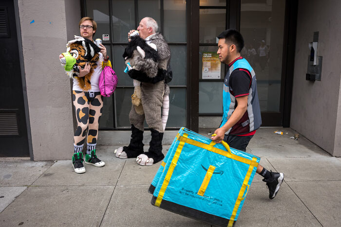 Street photographer captures humor and chaos with people in animal costumes and delivery worker on city sidewalk.
