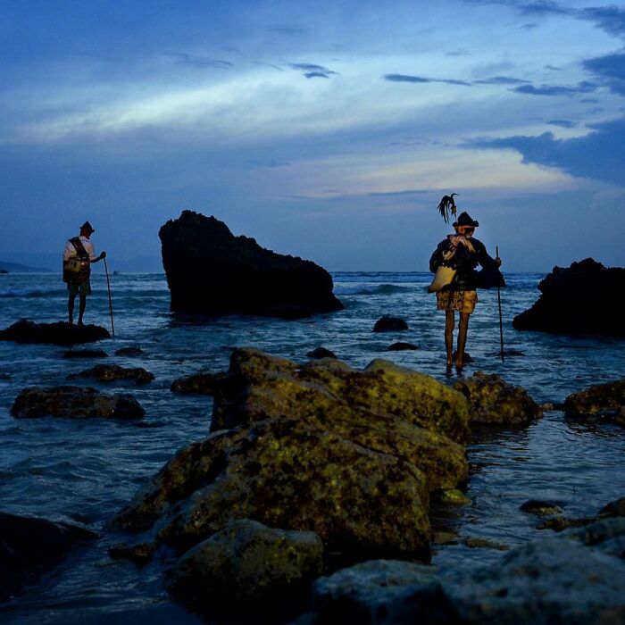 Two people standing on rocky shore at dusk, captured through Harjanto Sumawan’s candid photos of life’s beauty.