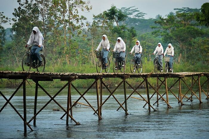 Students in white hijabs riding bicycles across a narrow wooden bridge, capturing life’s beauty through candid photos.