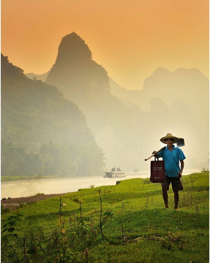 Man carrying farming tools on green riverside with mountains in background, captured through Harjanto Sumawan’s lens.