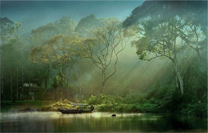 Misty morning scene captured through Harjanto Sumawan’s lens with a boat on calm water and sunlight filtering through trees.