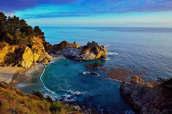 Scenic coastal view captured through Harjanto Sumawan’s lens showing ocean, rocks, trees, and clear blue sky at sunset.