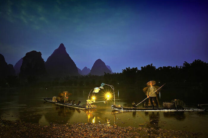 Fishermen on river at night with illuminated boats and mountainous landscape in candid photo capturing life’s beauty.