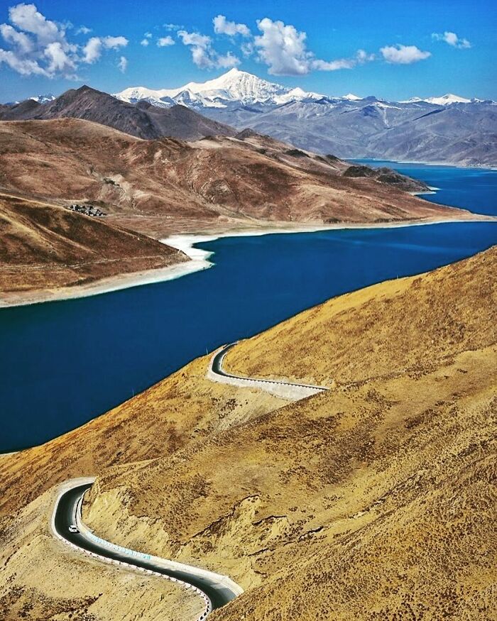 Winding road along a blue lake with barren hills and snow-capped mountains, showcasing candid photos capturing life’s beauty.