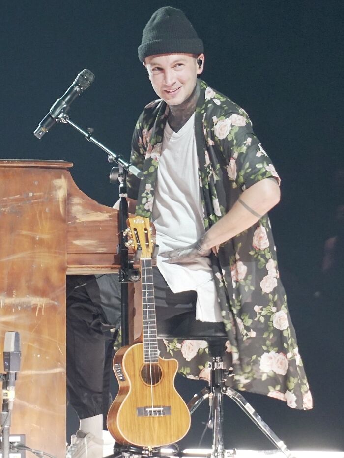 Musician wearing a floral shirt and beanie sitting by a piano with a ukulele during a live celebrity performance.