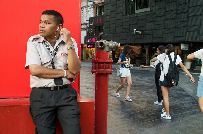 Man leaning against red wall with earphones as people walk by, showing street coincidences in everyday urban life.