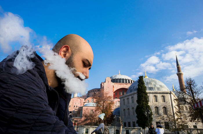 Man with smoke around his face creating a witty street coincidence near historic buildings under a blue sky.