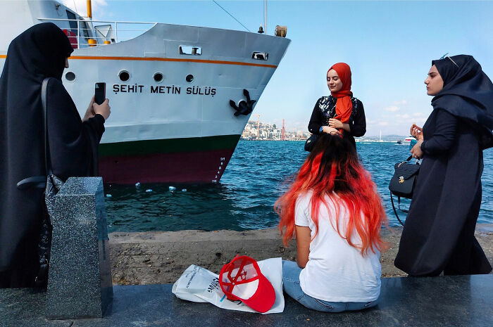 Four women by the water near a ship, one with red hair and another taking a photo, showcasing witty street coincidences.