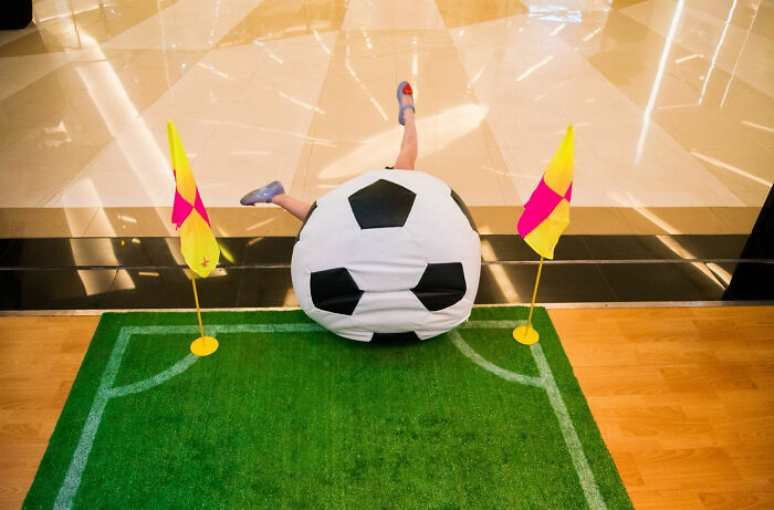 Person dressed as a giant soccer ball playfully falling on a small indoor soccer field in a witty street coincidence photograph.