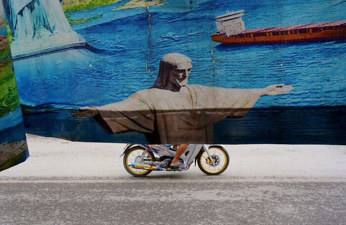 Motorbike rider appears to be part of street art with Jesus statue’s arms, showcasing witty street coincidences captured.