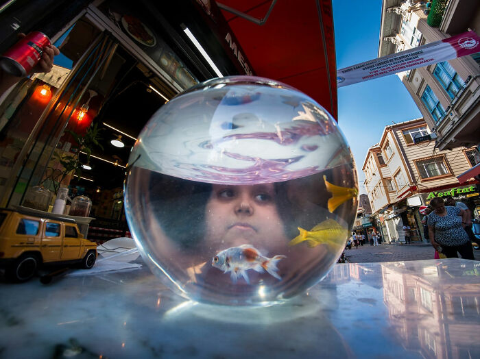 Child’s face distorted inside a fishbowl with goldfish, capturing a witty street coincidence on a busy city sidewalk.
