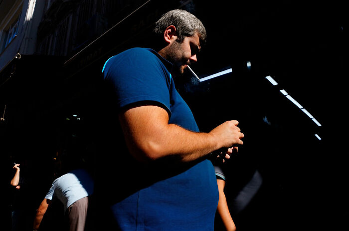 Man in a blue shirt smoking a cigarette with strong light and shadows in a street coincidence photo by Tavepong Pratoomwong.