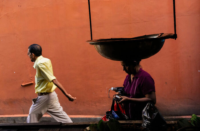Man walking past woman with large basin above her head, a witty street coincidence captured on a vibrant orange wall background.