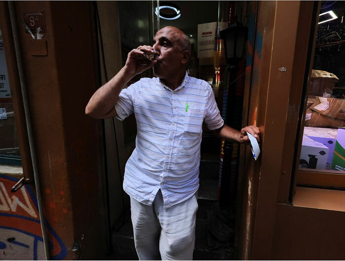 Man drinking tea while exiting a doorway, a candid street coincidence captured by Tavepong Pratoomwong.