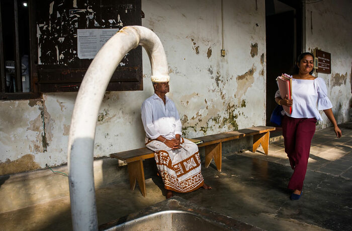Man sitting on a bench with a curved pipe aligning perfectly with his head in a witty street coincidence photo.