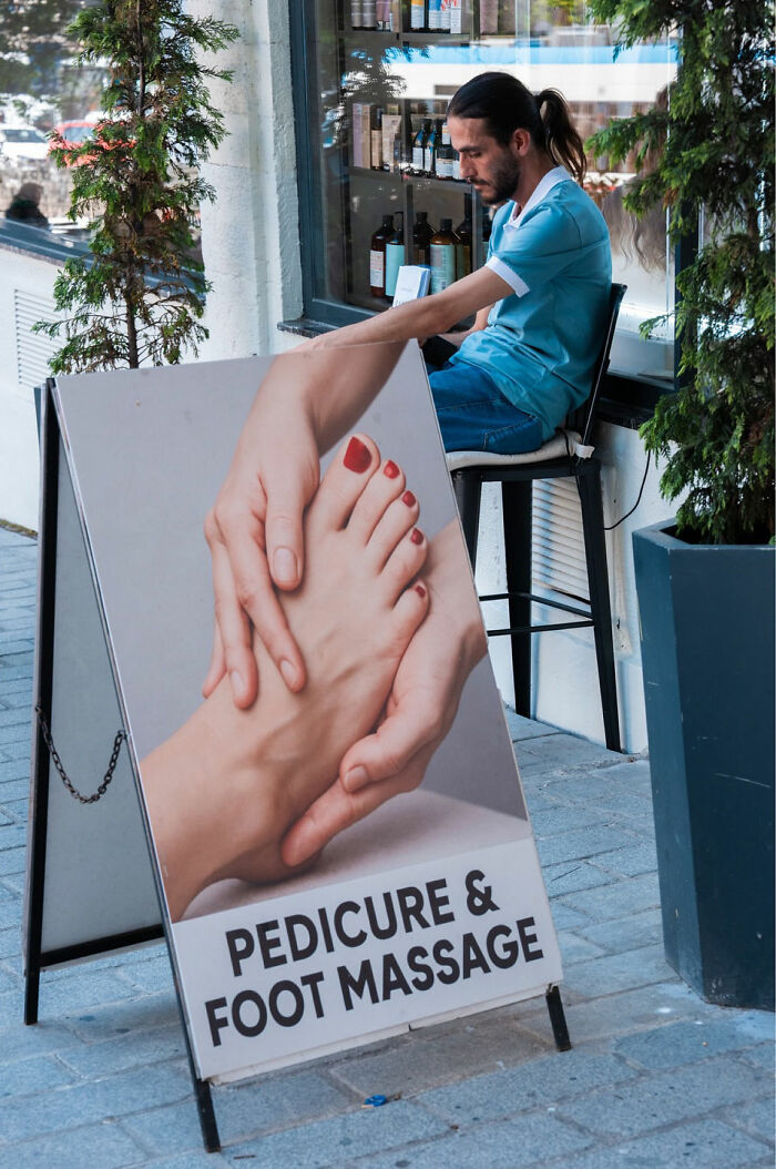 Man sitting behind a pedicure and foot massage sign creating a street coincidence captured by Tavepong Pratoomwong