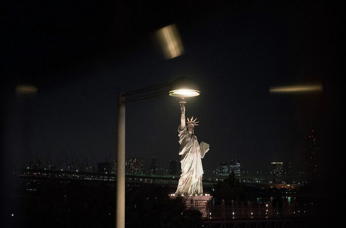 Street coincidences captured at night showing the Statue of Liberty illuminated by a street lamp against a city skyline.