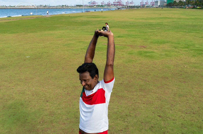 Man stretching arms on grassy field with a miniature person sitting on his hands in a witty street coincidence photo.
