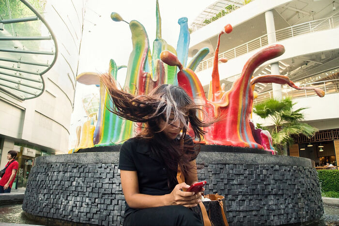 Woman sitting by colorful sculpture with hair blowing, captured in a captivating and witty street coincidence moment.
