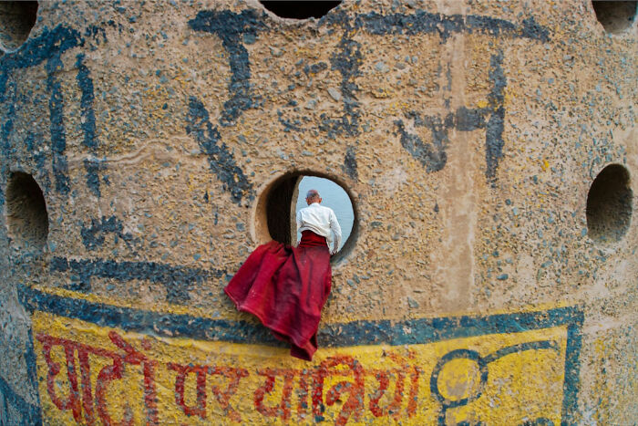 Man in red skirt perfectly aligned with round hole in textured wall, showing a witty street coincidence captured in urban setting.