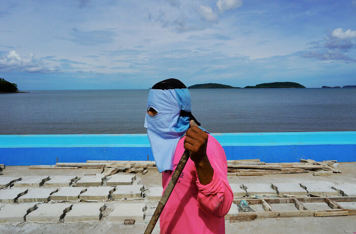 Man wearing a blue face cover and pink shirt holding a stick near the sea, capturing a street coincidence moment.