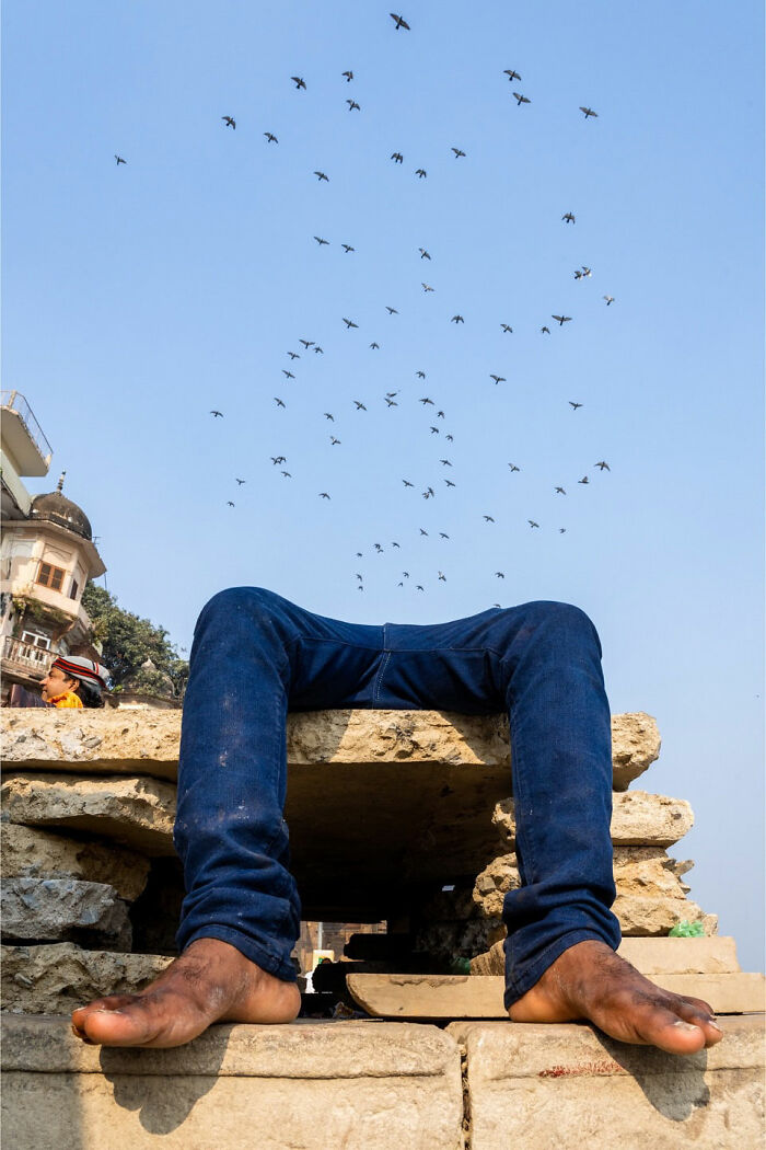 Man sitting on stone structure with legs visible and birds flying above in a captivating street coincidence photo