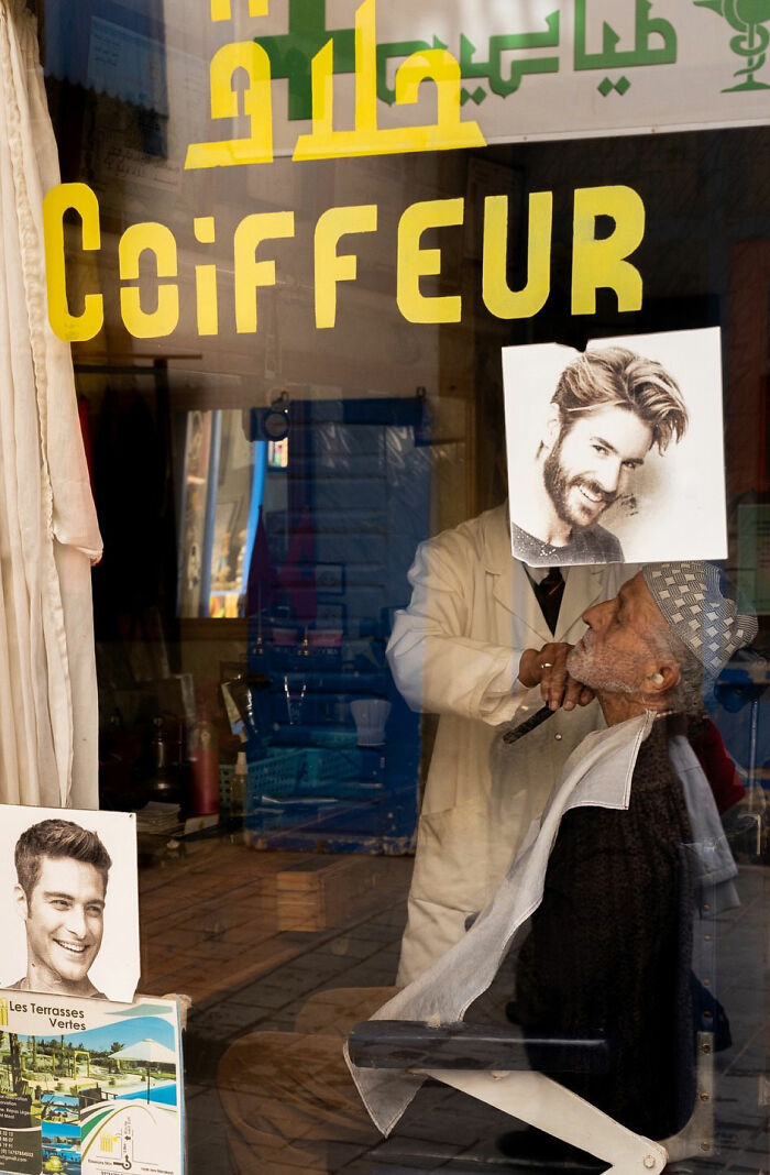Reflection shows barber matching haircut style with an elderly man in a street coincidence captured by Tavepong Pratoomwong.