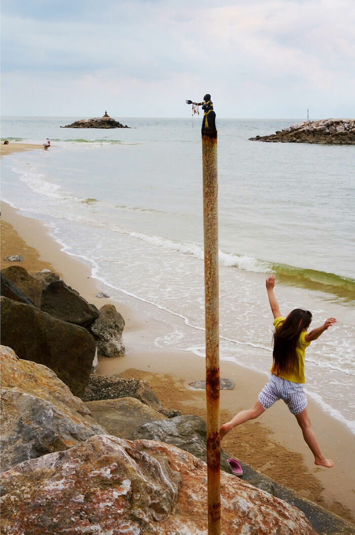 A child jumping on the beach near a pole with a sculpture, showcasing captivating street coincidences by Tavepong Pratoomwong.