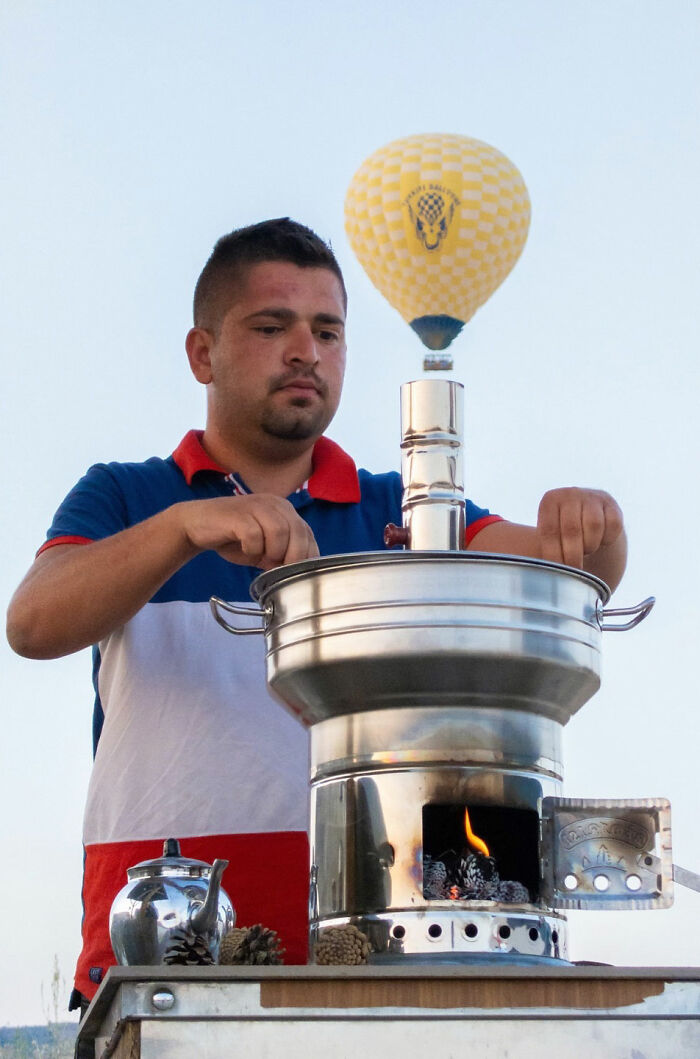 Man preparing tea on a street stove with a hot air balloon perfectly aligned above, showcasing a witty street coincidence.