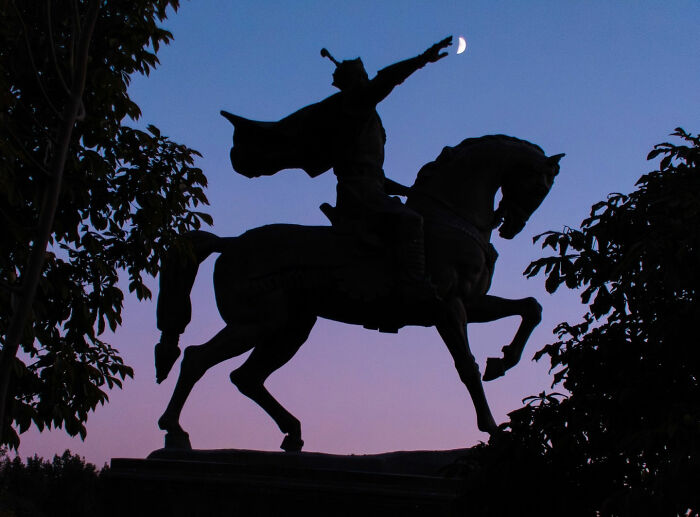 Silhouette of a street statue on horseback with a rider reaching toward the crescent moon in a captivating street coincidence.