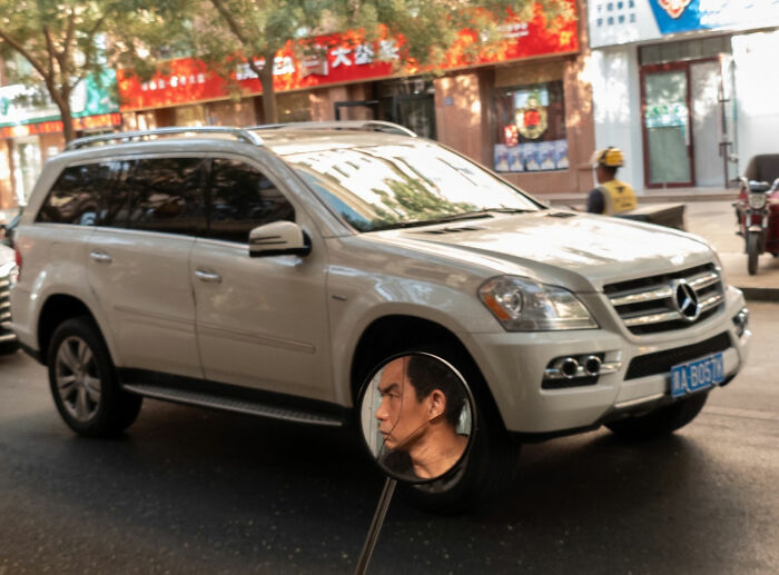 Silver Mercedes SUV parked on a city street with a man's face reflected in a nearby circular mirror, street coincidences.