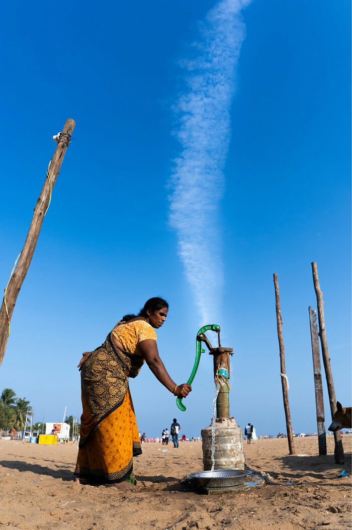 Woman in traditional attire using a hand pump with water flowing, capturing a witty street coincidence moment outdoors.