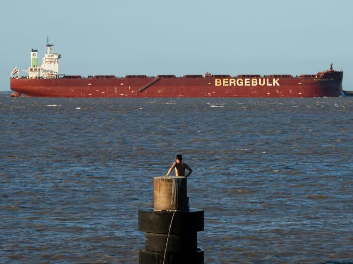 Person leaning on a post by the water with a large cargo ship in the background, a captivating street coincidence.