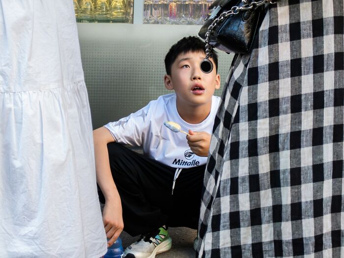 Young boy squatting on the street with a spoon, surrounded by pedestrians in a candid street coincidence moment.